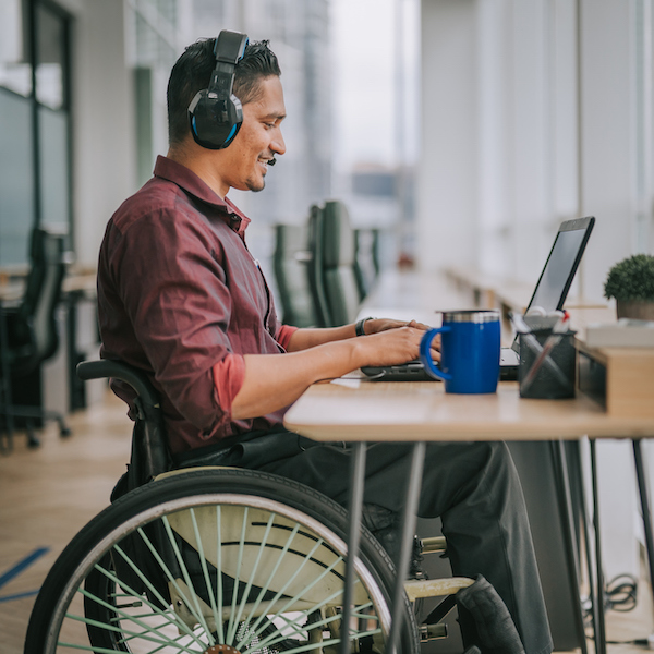 A man in a wheel chair wearing headphones working on his laptop in an office setting.