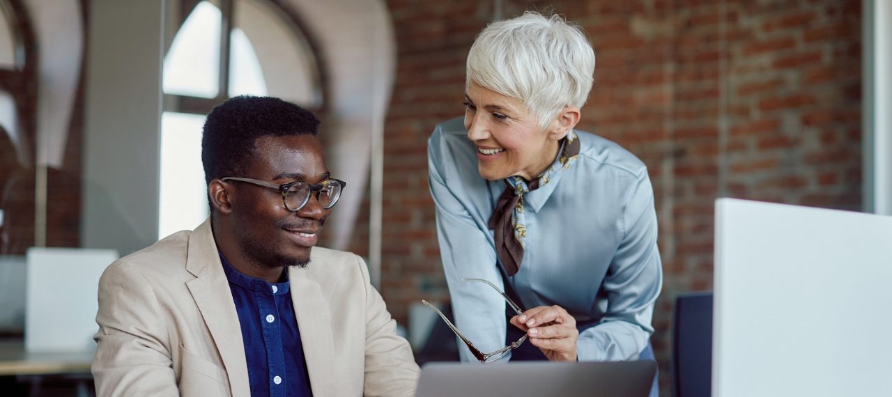 A smiling older woman with short white hair leans over to engage in conversation with a younger man wearing glasses and a beige blazer. They are in a modern office setting with exposed brick walls, natural light, and a laptop open in front of them. The image conveys a collaborative and inclusive work environment, reinforcing the theme of disability inclusion in hiring.